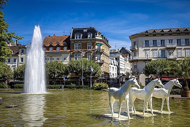 Springbrunnen auf dem Augustaplatz in Baden-Baden, links im Vordergrund eine Skulpturengruppe von drei weißen Pferden.